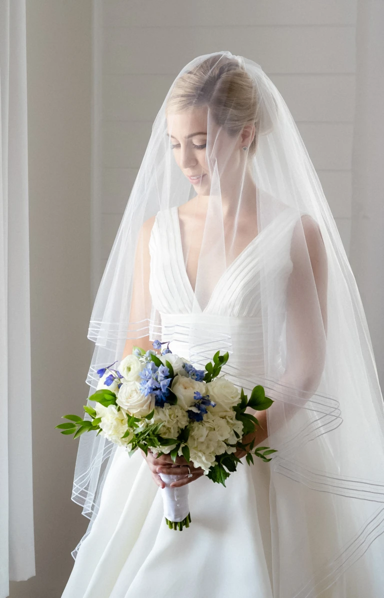 Bride in a sheer veil holding a white and blue bouquet by a window