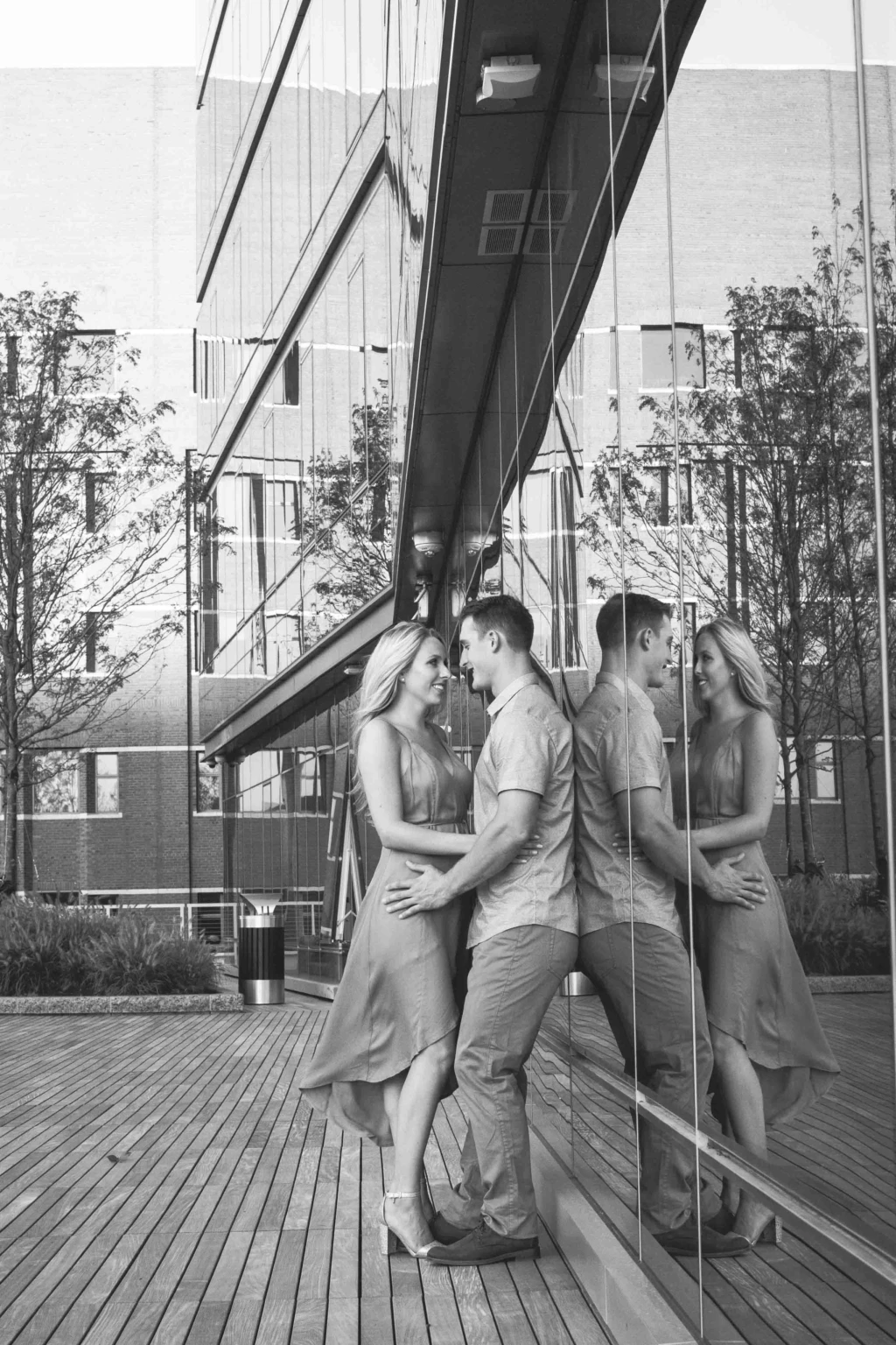 Black-and-white engagement portrait of a couple reflected in a glass building
